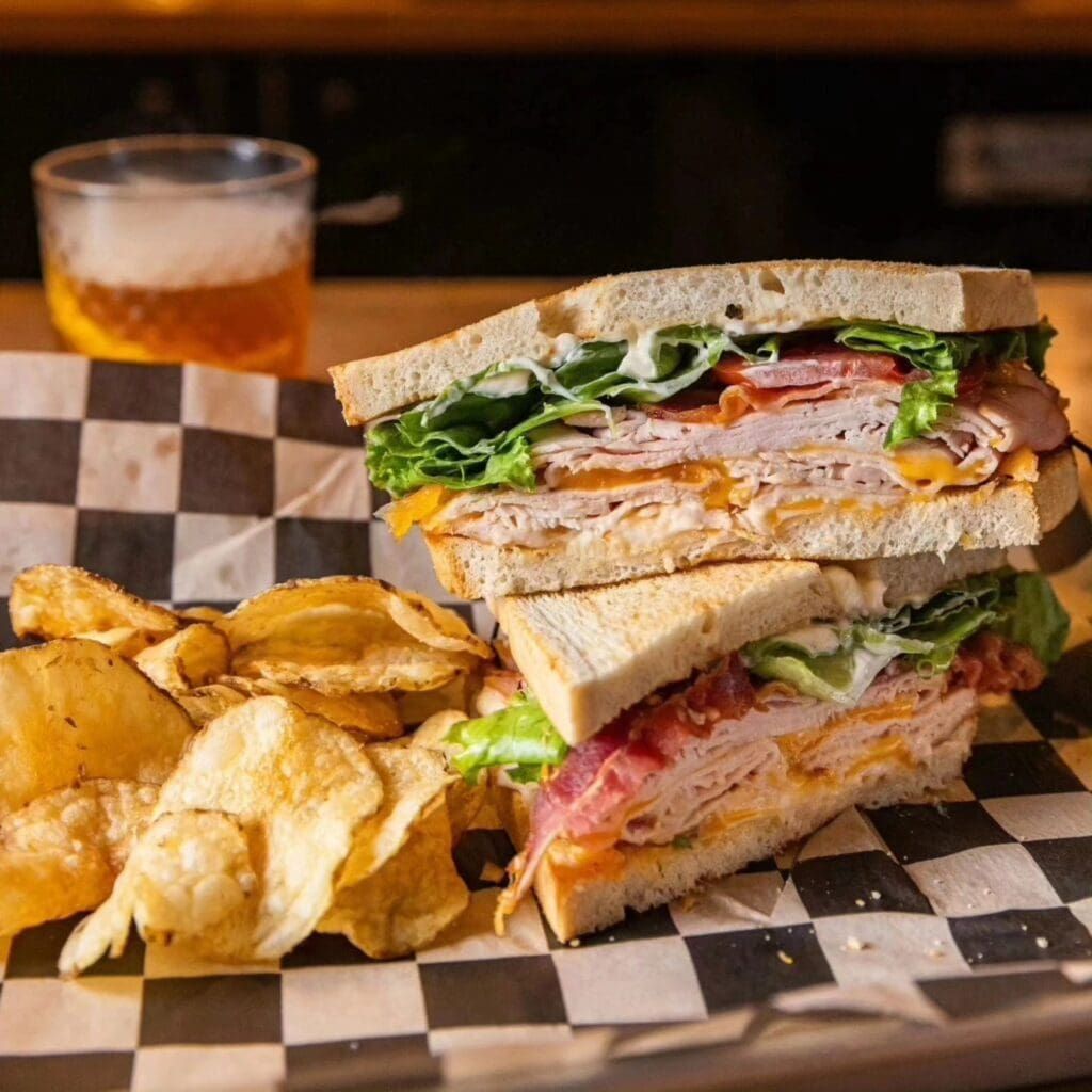Two stacked deli sandwiches with meats, greens, and cheese served with chips
