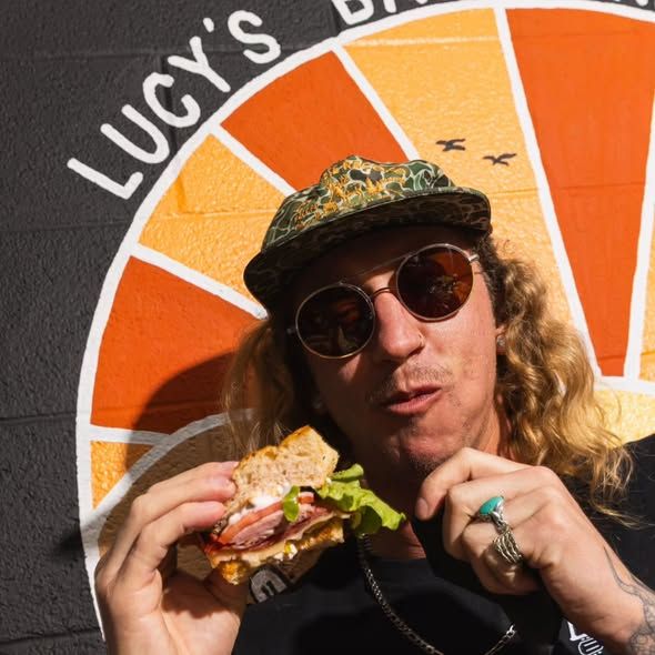 A person in sunglasses and a hat holding a substantial sandwich in front of Lucy's orange and yellow signage