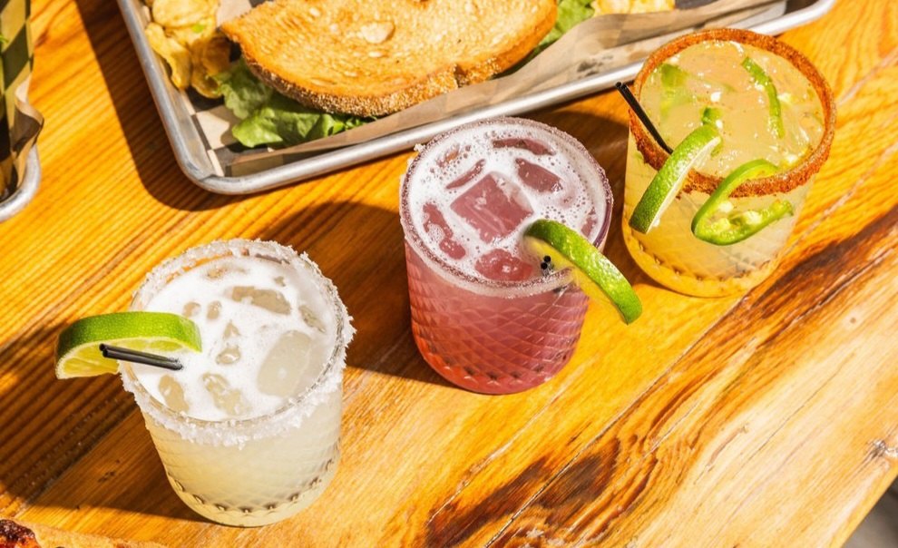 Three margaritas with salted rims and lime wedges on a sunlit wooden table