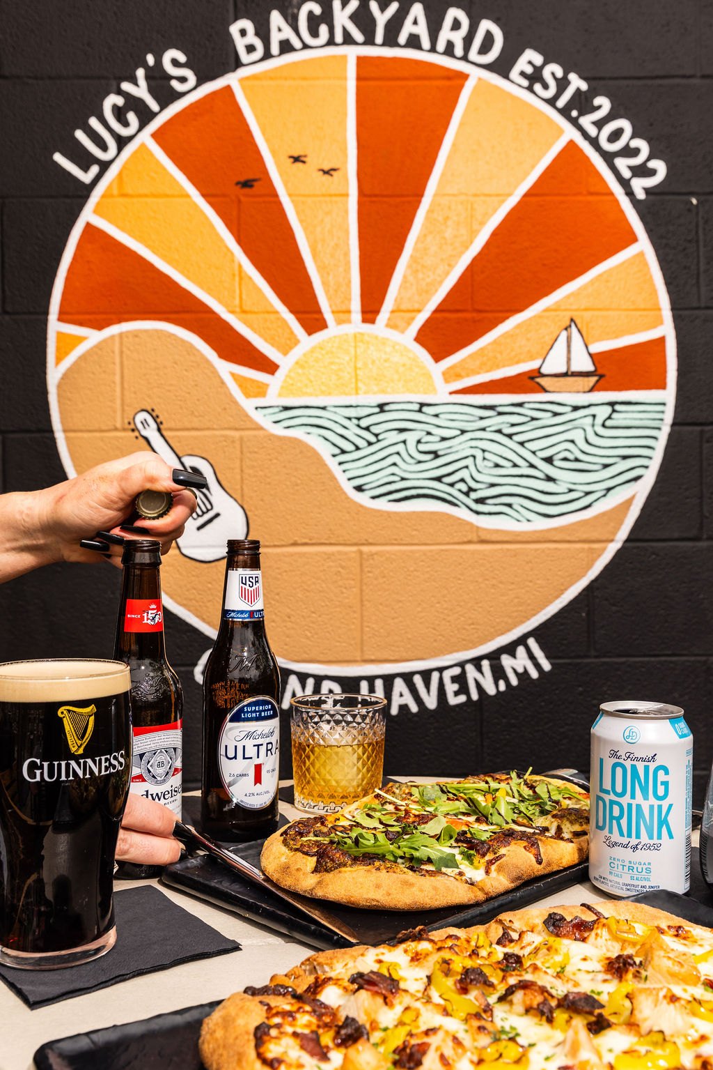 Bottles, cans and a stout in front of the Lucy's Backyard sunrise mural