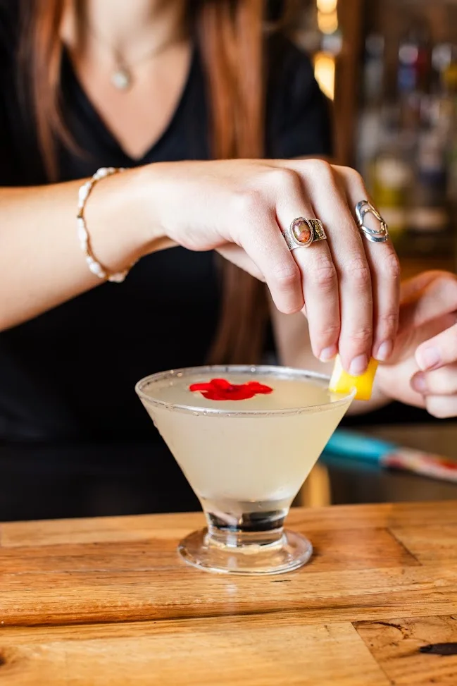 A bartender adding a lemon twist to a martini topped with a red flower petal