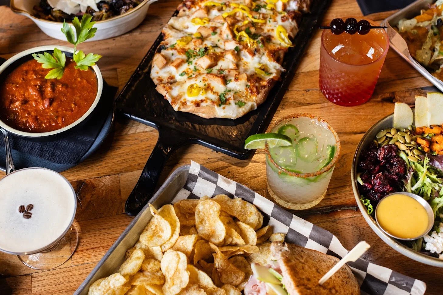 A bowl of chili with a parsley garnish beside a chicken flatbread, a margarita, an espresso martini, chips with a sandwich, and a salad