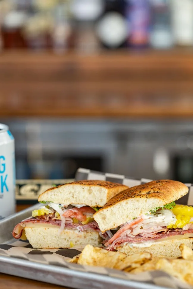 An Italian-style sandwich with salami, capicola and banana peppers on focaccia, cut in half, with chips and a can beside it