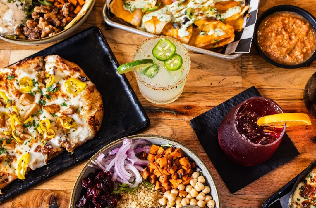 A table from above with a chicken and banana pepper flatbread, a jalapeño margarita, a tray of loaded nachos with salsa, a grain bowl and a berry cocktail