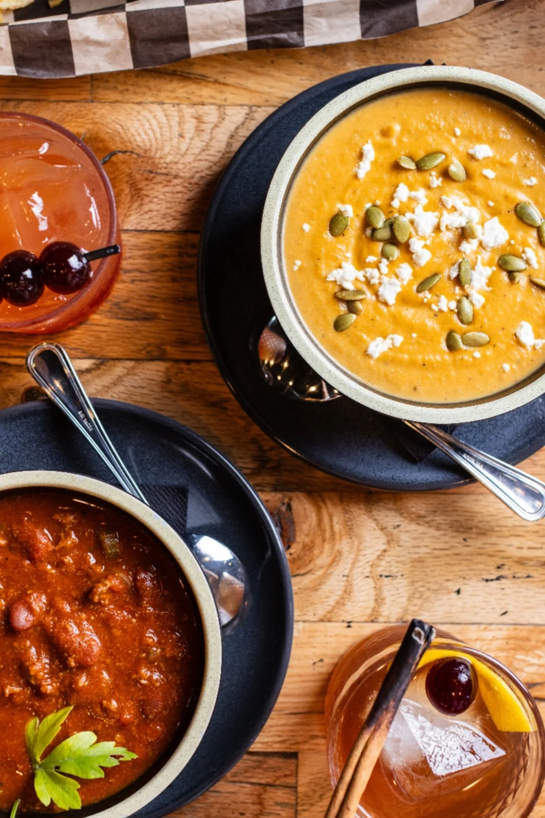 A bowl of creamy orange soup topped with feta and pumpkin seeds beside a bowl of chili, with two cocktails on a wooden table