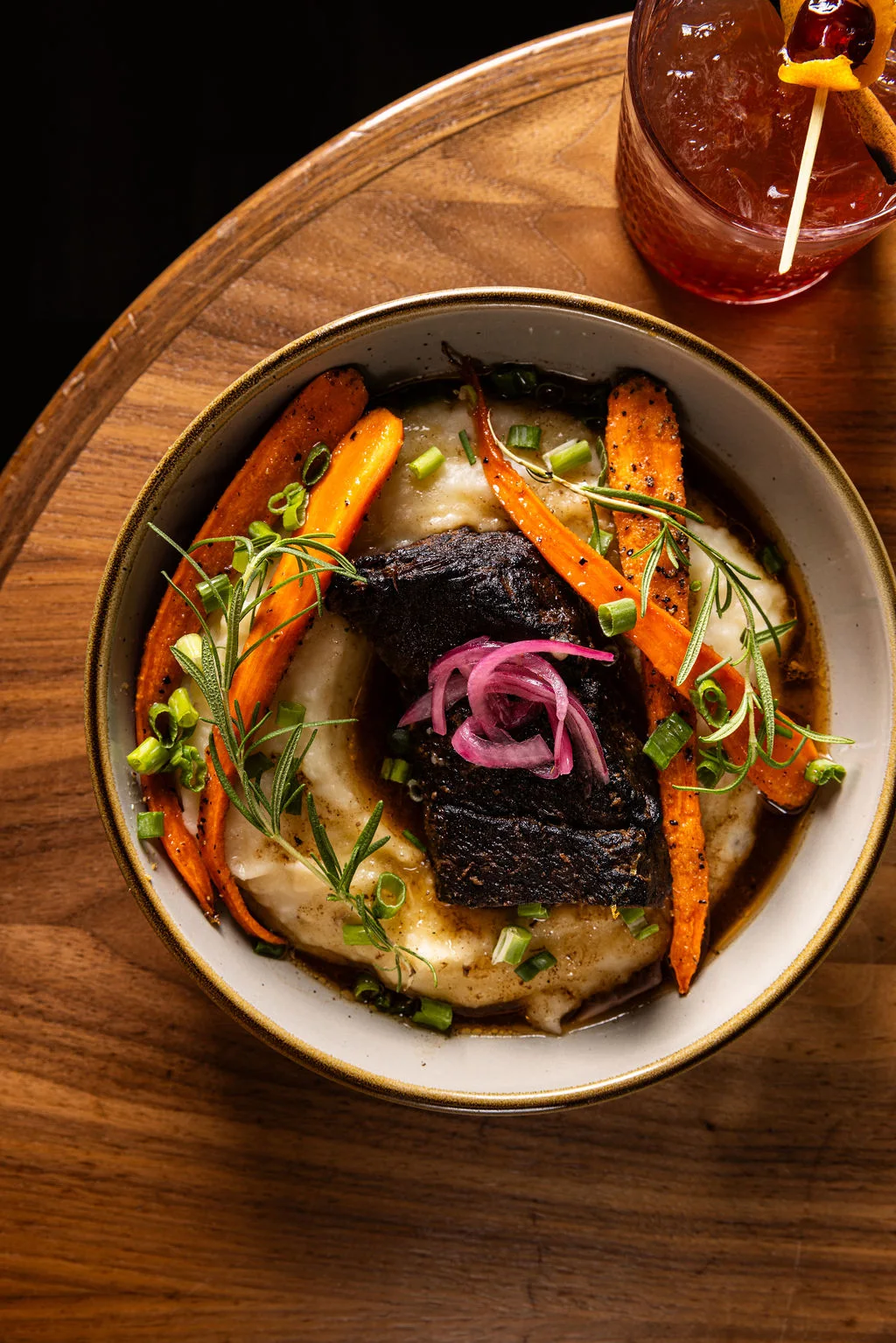 A bowl of braised short rib over creamy mashed potatoes with roasted carrots, rosemary and pickled red onion, with a cocktail beside it