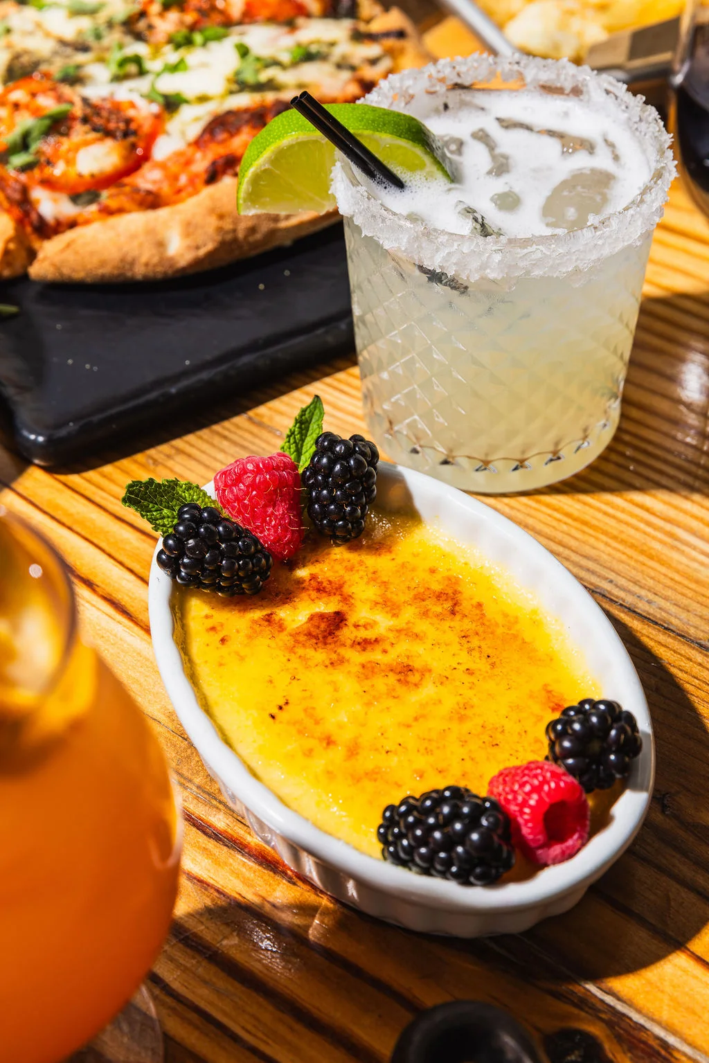 A salt-rimmed margarita in the sun beside a flatbread and a crème brûlée topped with berries